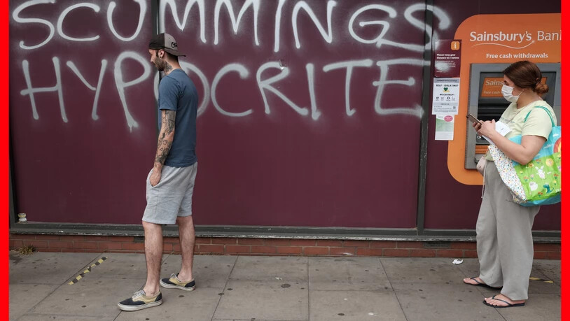 Ein Mann und eine Frau mit Mundschutz gehen an einem Graffiti vorbei, das an die Wand eines Supermarkts in der Nähe des Hauses von Regierungsberater Cummings gesprüht wurde. Das Graffiti mit der Aufschrift "Scummings Hypocrite" (Heuchler)  kritisiert…