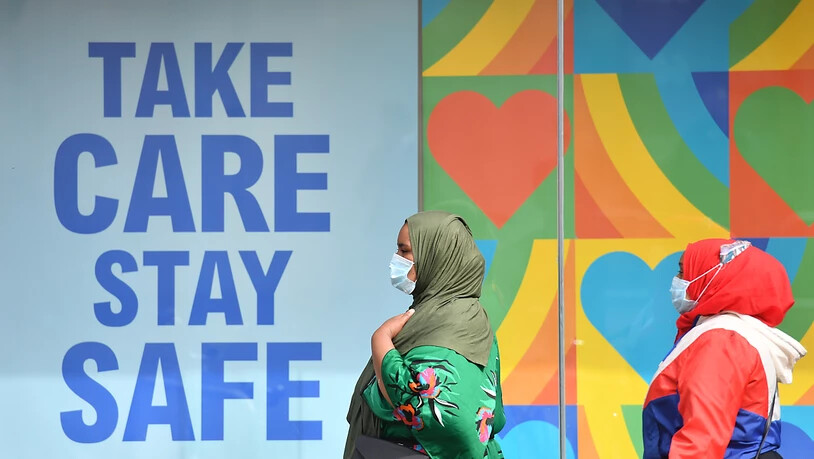 Frauen mit Kopftuch und Mundschutz gehen in einem Stadtzentrum in Großbritannien an einem Plakat vorbei, das an die Corona-Schutzregeln erinnert: "Take care, stay safe". Foto: Jacob King/PA Wire/dpa